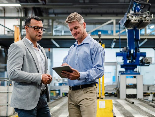 Two men in a factory setting discuss something while looking at a tablet. Machinery and industrial equipment are visible in the background.