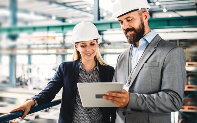 Two people in business attire and hard hats stand in an industrial setting, discussing information on a tablet.