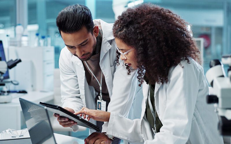 Two scientists in lab coats are examining a tablet screen together in a laboratory setting with microscopes and equipment nearby.