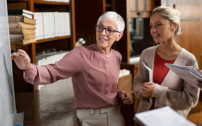 Two women discussing in a library, with one writing on a whiteboard and the other holding documents.