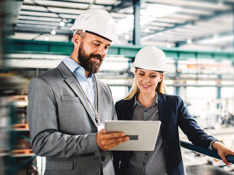 Two people in hard hats view a tablet in an industrial setting. The man wears a suit, and the woman is in business attire.