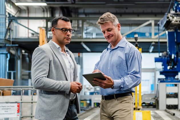 Two men in a factory setting discuss something while looking at a tablet. One wears a suit jacket, the other a light blue shirt. Machinery and equipment are visible in the background.