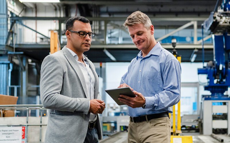 Two men in a factory setting discuss something while looking at a tablet. One wears a suit jacket, the other a light blue shirt. Machinery and equipment are visible in the background.