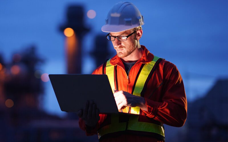 A worker in a hard hat and safety vest uses a laptop at an industrial site during twilight.