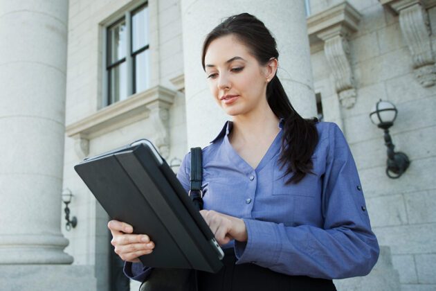 A woman in a blue shirt stands outside a building, using a tablet.