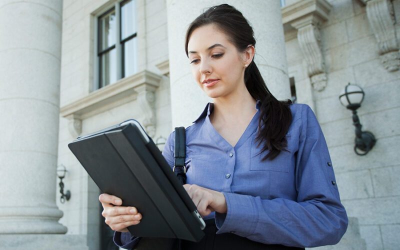 A woman in a blue shirt stands outside a building, using a tablet.