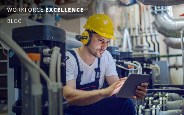 Worker in overalls and a yellow helmet uses a tablet in an industrial setting with machinery visible in the background.