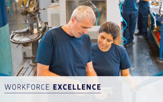 Two factory workers in navy shirts look at a tablet together in an industrial workspace. Text overlay reads "WORKFORCE EXCELLENCE powered by transparent and accurate pay.