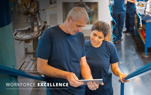 Two employees in blue uniforms review a tablet in an industrial setting, ensuring transparent and accurate pay for workforce excellence.