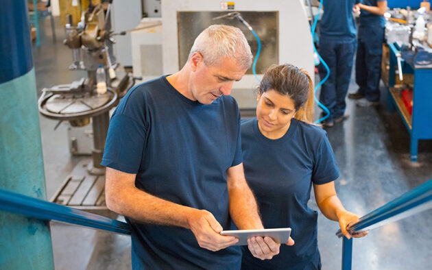 Two people in a workshop looking at a tablet, with machinery in the background. Both are wearing blue shirts.