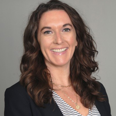 A woman with long brown wavy hair, wearing a black blazer and a striped shirt, smiles against a plain background.