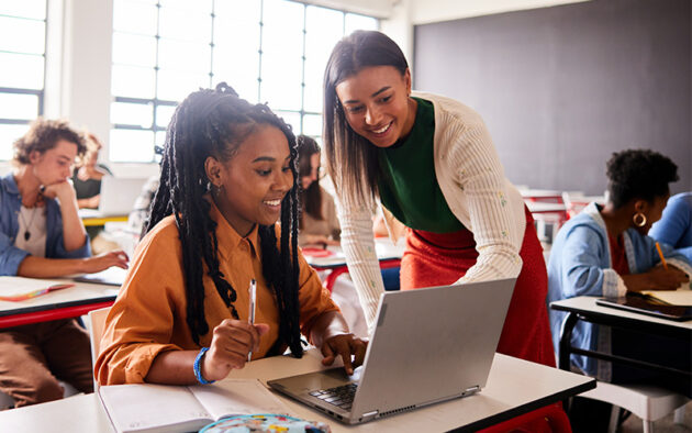 A teacher assists a student working on a laptop in a classroom, while other students are focused on their own work in the background.