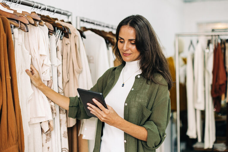 A woman stands by racks of clothing, holding and using a tablet, wearing a white top and green shirt in a clothing store.