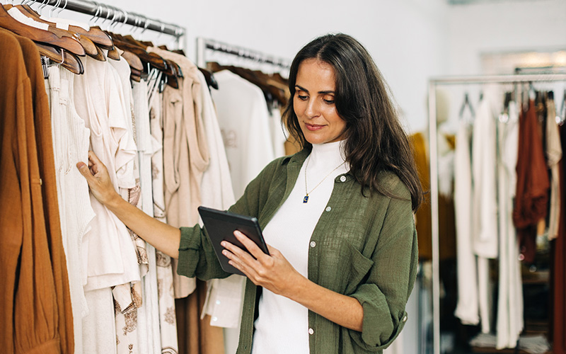 A woman stands between clothing racks, holding a tablet and examining garments in a boutique or store setting.