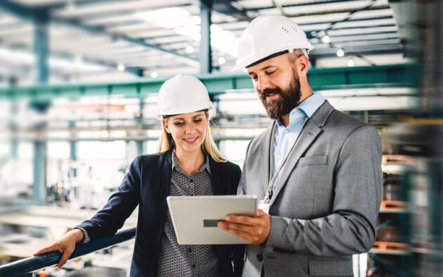 Two people in business attire and hard hats review a tablet inside an industrial facility.