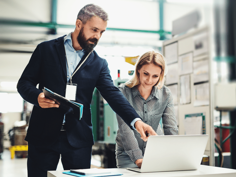 Two people in a professional setting, one standing and pointing at a laptop while the other observes. They're surrounded by office equipment and papers.