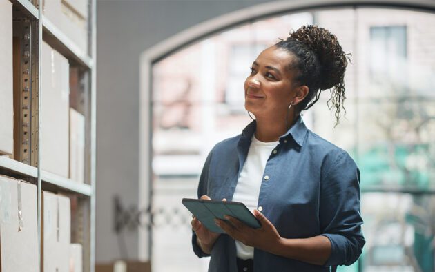 A woman in a blue shirt stands in a warehouse, holding a tablet and looking at shelves with a thoughtful expression.