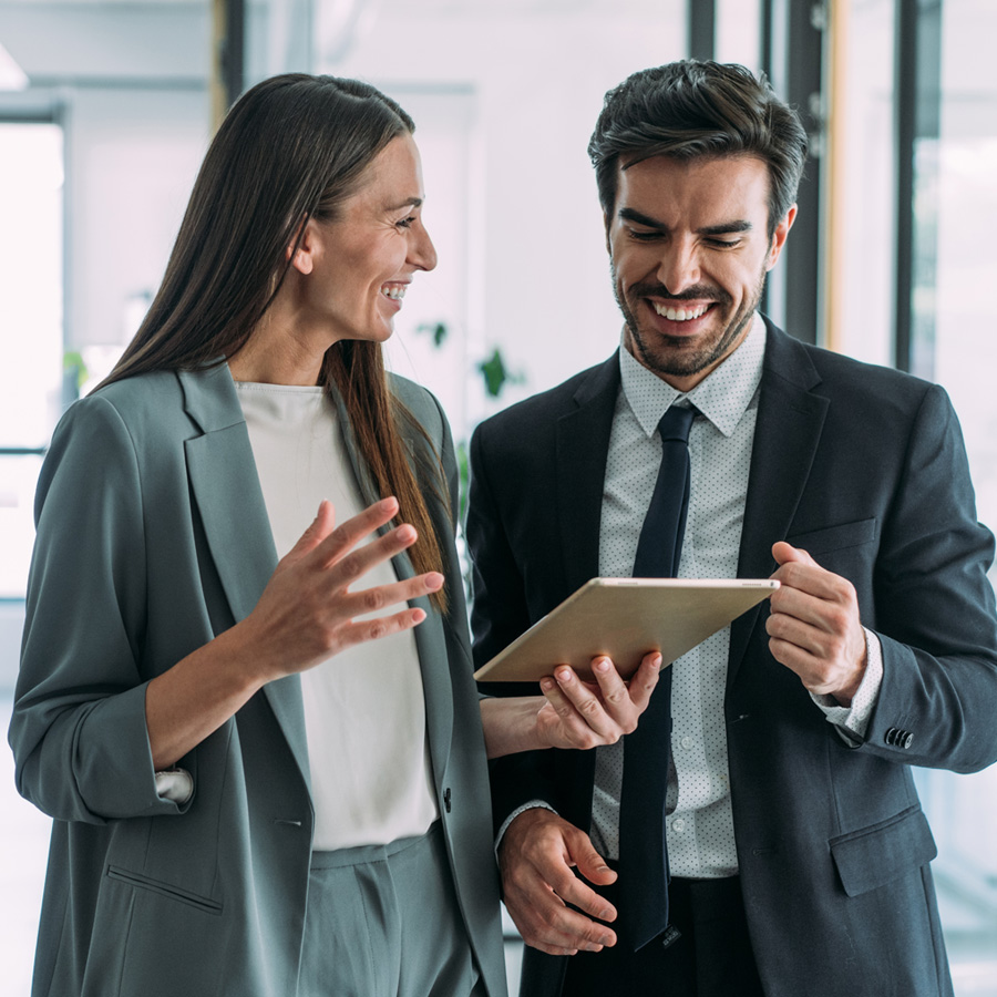 Two business professionals smiling and looking at a tablet together in an office setting.