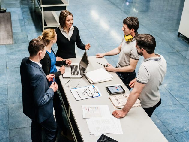Five people in a business setting stand around a table with laptops and documents, having a discussion.