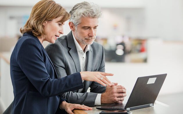 Two business professionals, a woman and a man, are sitting at a desk, focused on a laptop screen in an office setting.