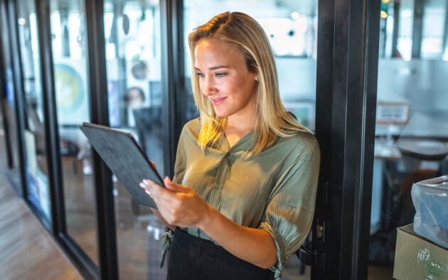 A woman in a green shirt stands in an office, looking at a tablet and smiling. She leans against a glass wall with wooden frames, surrounded by office furniture and equipment.