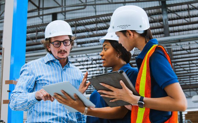 Three individuals wearing hard hats and holding tablets discuss in an industrial setting. One person wears a safety vest.