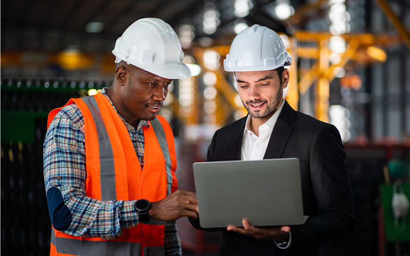 Two men wearing hard hats, one in a safety vest and the other in a suit, look at a laptop in an industrial setting.