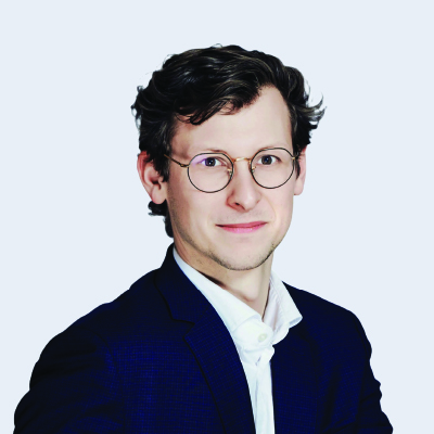 A man with short wavy hair and glasses, wearing a dark suit jacket and white shirt, poses against a plain light background.