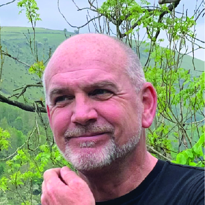 A middle-aged man with a gray beard and short hair stands outdoors, surrounded by green trees and hills, wearing a black shirt.
