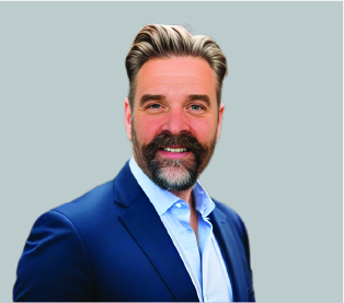 A man with styled hair, a beard, and mustache, wearing a blue suit jacket and light blue shirt, poses and smiles in front of a plain light gray background.
