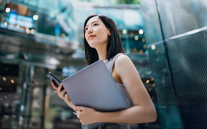 A woman holding a laptop and smartphone stands in a modern glass building, looking ahead with a neutral expression.