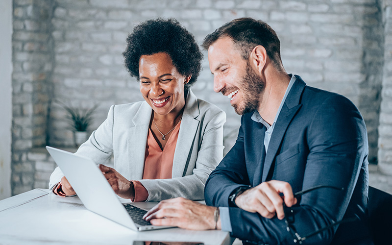 Two professionally dressed people sit at a table, smiling and looking at a laptop screen together in a modern office setting.