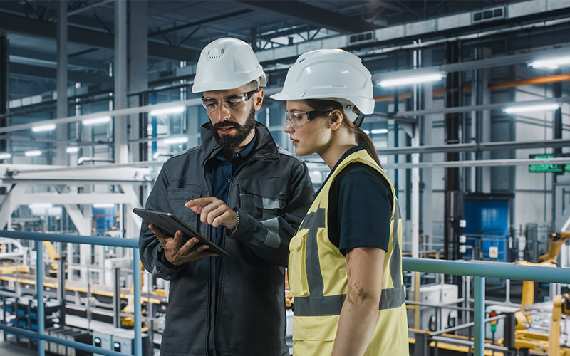 Two workers wearing hard hats and safety glasses stand in a factory, reviewing information on a tablet. Industrial equipment and metal structures are visible in the background.