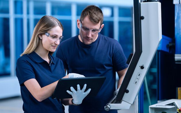Two people wearing safety glasses and navy shirts review information on a tablet in an industrial or manufacturing setting.