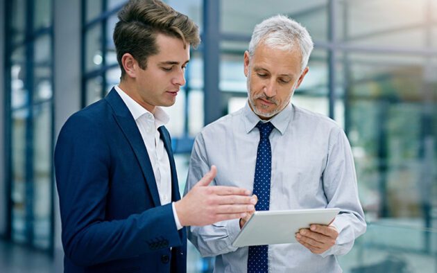 Two men in business attire stand indoors, looking at a tablet. One man gestures while speaking, and the other listens attentively, holding the device.