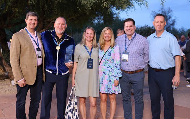 Six people stand outdoors, posing for a group photo; one holds an award plaque and another wears a medal. They are dressed in business casual attire, smiling at the camera.