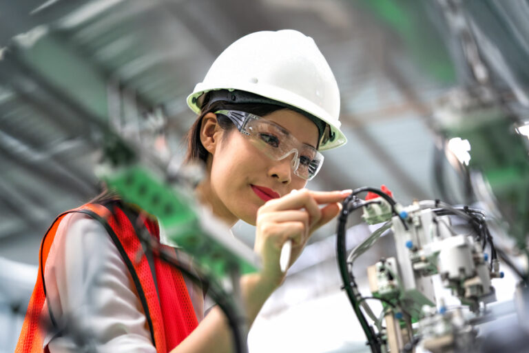 A woman wearing a white hard hat, safety glasses, and an orange vest inspects and adjusts machinery in an industrial setting.