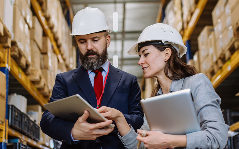 Two people wearing suits and safety helmets review information on a tablet in a warehouse filled with shelves and boxes.