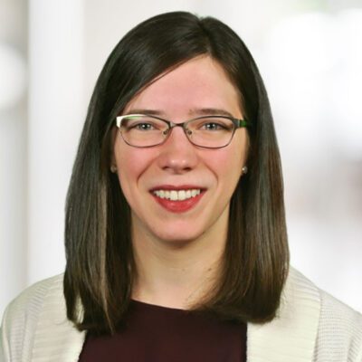 A woman with straight brown hair, glasses, and a white sweater smiles at the camera against a blurred indoor background.