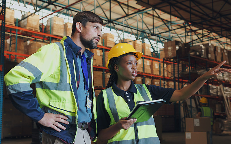 Two warehouse workers in safety vests and helmets stand inside a warehouse; one holds a tablet while gesturing, the other observes the direction indicated.