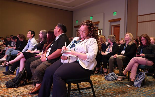 People seated in rows at a conference or seminar, with some taking notes and others listening attentively.