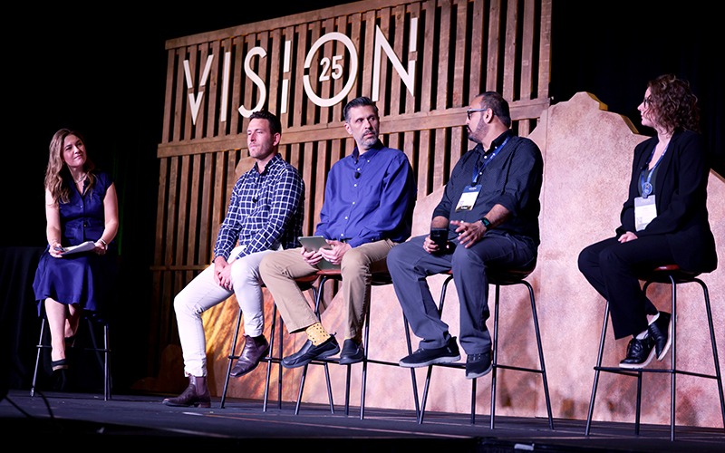 Five people sit on high stools on a stage, engaged in a panel discussion in front of a "VISION 25" sign.