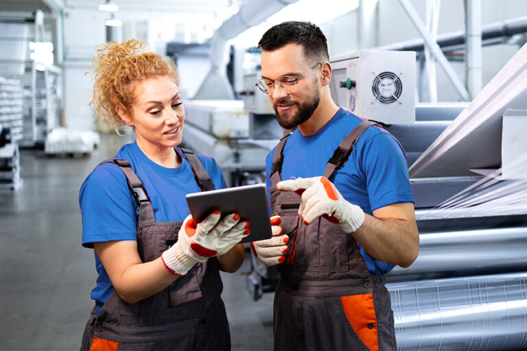 Two factory workers in blue shirts and overalls review information on a digital tablet while standing in an industrial setting with machines in the background.