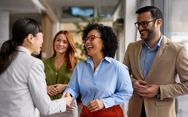 Four professionally dressed people stand indoors, smiling and talking, with two women in the foreground shaking hands.