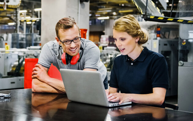 Two people in work uniforms look at a laptop together at an industrial facility, appearing to discuss something on the screen. Machinery is visible in the background.
