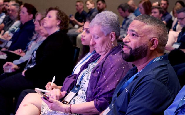 A group of people seated in an audience, some taking notes and listening attentively at what appears to be a conference or seminar.
