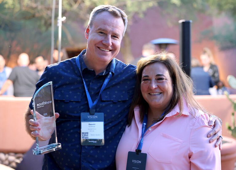 A man holding an award trophy stands next to a smiling woman. Both are wearing conference name badges and casual clothing at an outdoor event.