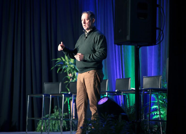 A man stands on a stage giving a presentation, with several empty chairs, plants, and dark curtains in the background.