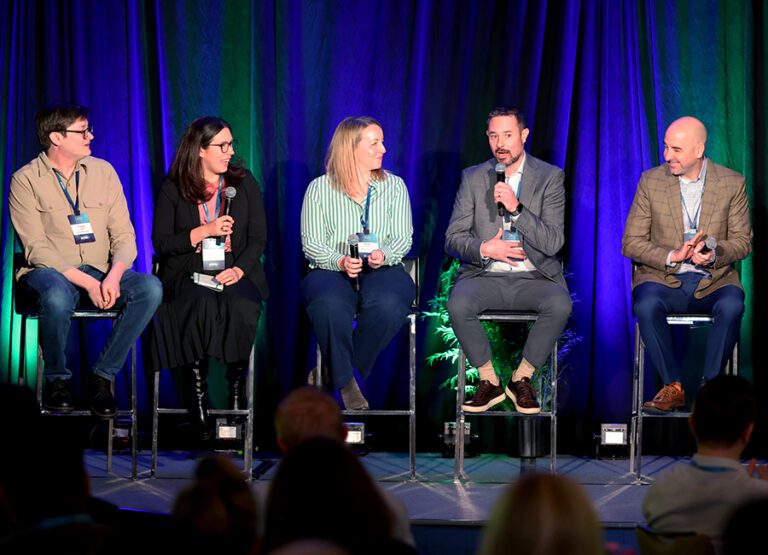 Five people sit on high stools on stage during a panel discussion, each holding a microphone, with a dark curtain backdrop and audience members visible in the foreground.