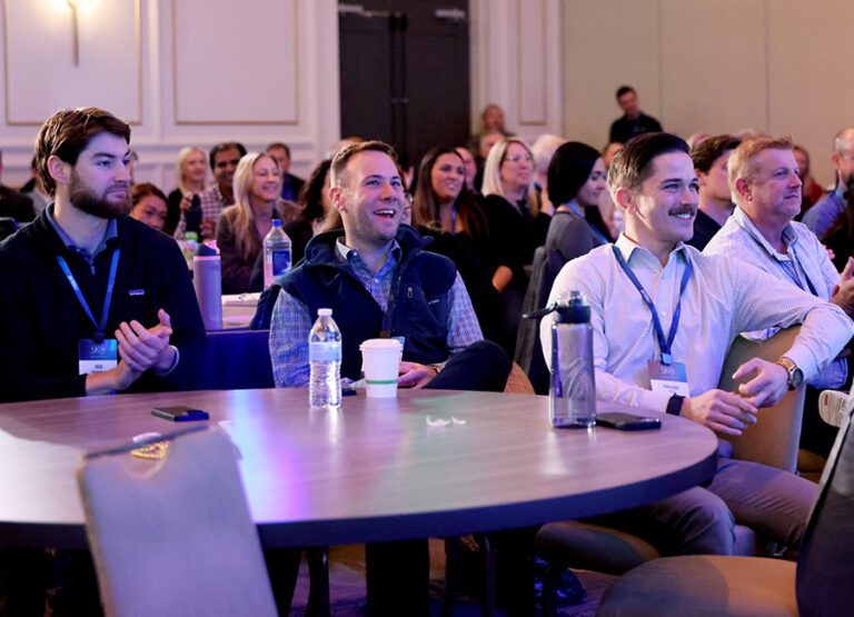 A group of people sit at round tables in a conference room, attentively watching a presentation and smiling. Bottles, cups, and papers are on the tables.